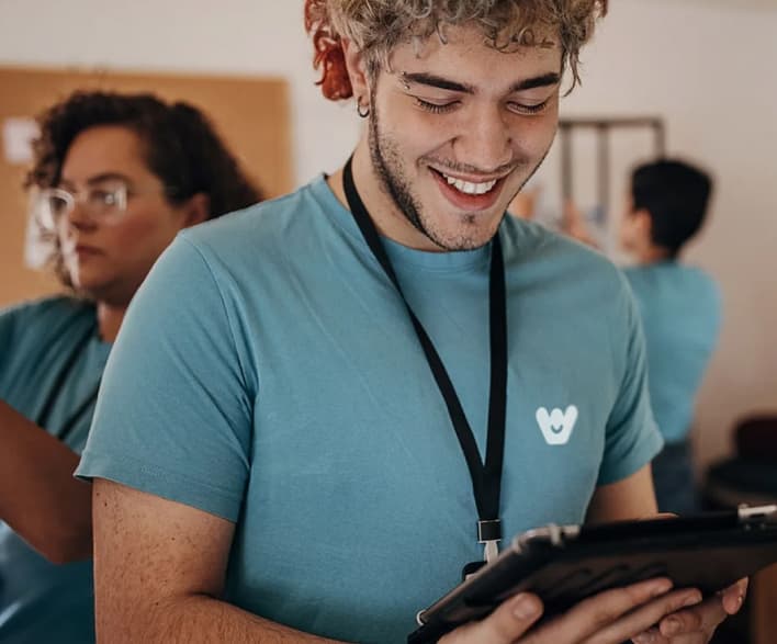 Un giovane volontario con i capelli ricci e sfumature rosse sorride mentre usa un tablet. Indossa una maglietta azzurra con il logo di Wishraiser e un badge al collo. Sullo sfondo, altri volontari lavorano in un ambiente collaborativo